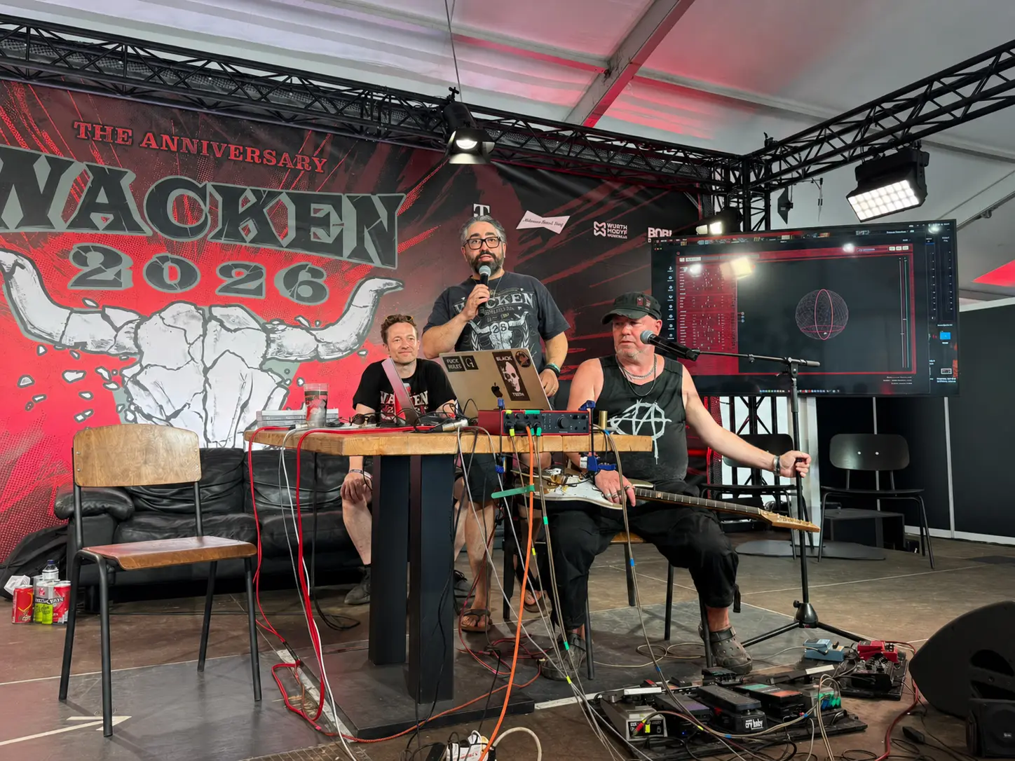 Press conference at Wacken Open Air 2026: Christian Stein and Bob Coecke seated at a table, Manouchehr Shamsrizi standing with a microphone. Bob Coecke holds the quantum guitar; a large screen behind them shows the live quantum visualisation.